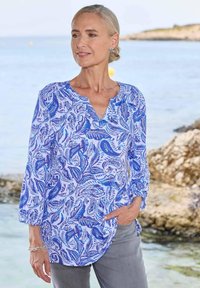 Langsleeve blouse in lichtblauw en wit met een paisleypatroon, voorzien van een V-hals en geraffineerde mouwen, gedragen met grijze pantalon.