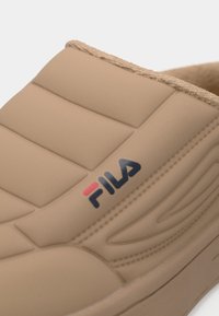 Beige polstret slip-on sko med myk foring og FILA-logo på siden, som viser detaljert søm og teksturert design.
