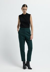 Top nero senza maniche con un motivo testurizzato e scollatura volant abbinato a pantaloni a vita alta in verde scuro a sigaretta e stivali neri con tacco.