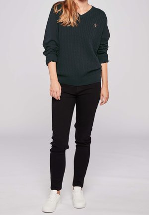 Kvinde iført mørkegrøn kabelstrikket sweater, sorte slim bukser og hvide sneakers, der står mod en neutral baggrund.