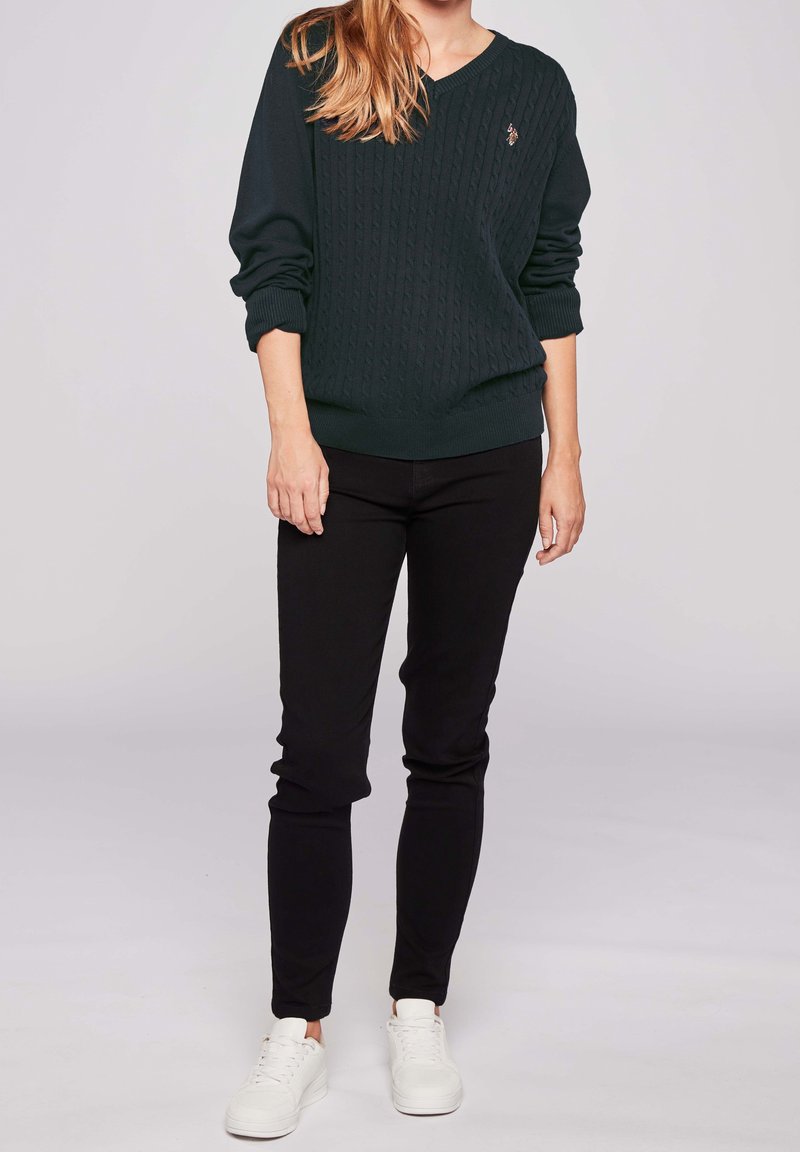 Femme portant un pull torsadé vert foncé, un pantalon slim noir et des baskets blanches, debout devant un fond neutre.