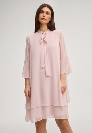 Kobieta ubrana w jasnoróżową, luźną sukienkę z warstwowym dołem, długimi, przejrzystymi rękawami oraz zawiązaną kokardą przy dekolcie, stojąca na jednolitym tle.
