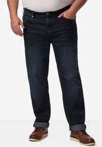 Jeans de mezclilla azul oscuro con un corte relajado, cierre de botón y bolsillos frontales. Se pueden ver los puños doblados y unos zapatos de cuero marrón resistentes.