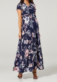 Pregnant woman wearing a navy blue floral wrap maxi dress with short sleeves and black open-toe heels, standing against a plain background.