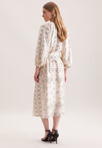Langes, cremefarbenes Kleid mit schwarzem Paisleymuster, Puffärmeln, gebundenem Taillengurt und Midi-Länge. Kombiniert mit schwarzen, hochhackigen Sandalen.