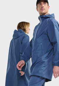 Hombre y mujer con chaquetas impermeables azules a juego con capuchas, de pie y mirando en direcciones opuestas sobre un fondo liso.