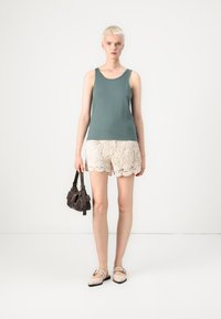 Tanktop i teal, blonde shorts i creme med bølger kant, og beige flade sko med stropdetaljer. Modellen bærer en lille mørk håndtaske.