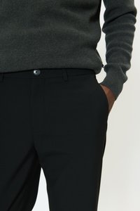 Vyras dėvi tamsiai pilką megztinį, įkištą į juodas pritaikytas kelnes, dešinė ranka kišenėje, stovi prie šviesios foninės sienos.