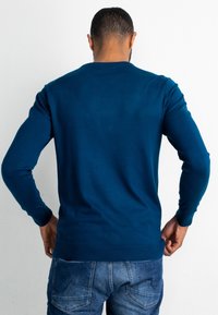 Dunkelgrüner, gestrickter Pullover mit Rundhalsausschnitt und langen Ärmeln, mit glatter Textur und geripptem Saum, kombiniert mit blauer Jeans.