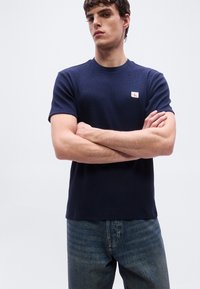 Νεαρός άντρας που φοράει μπλε πουκάμισο με κοντά μανίκια και ένα μικρό κόκκινο και λευκό λογότυπο, με σταυρωμένα τα χέρια, στέκεται μπροστά σε απλό λευκό φόντο.