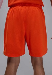 Oransje atletiske shorts laget av pustende meshstoff, med elastisk liv og perforert tekstur for bedre ventilasjon.