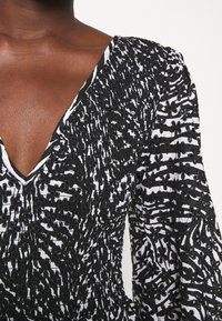 Černobílá vzorovaná blůza s výstřihem do V; texturovaná tkanina; dlouhé rukávy se skládáním; květinové akcenty po celé ploše.