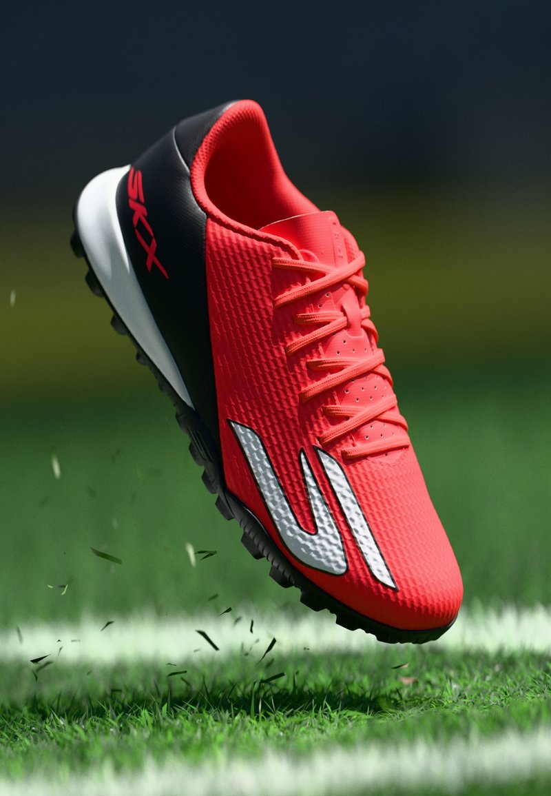 Φωτεινό κόκκινο ποδοσφαιρικό παπούτσι με μαύρη φτέρνα και λευκή σόλα, με το εμπορικό σήμα "SKX", αιωρείται πάνω από το γρασίδι με μικρά φύλλα να πετούν.