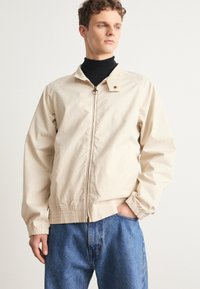 Hellbeige Jacke mit Reißverschluss und rundem Ziehgriff, gerippte Bündchen und Saum. Getragen über einem schwarzen Rollkragenpullover, kombiniert mit blauen Jeans.
