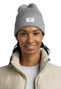 Gorro gris de canalé hecho de tejido de punto con una etiqueta que lleva el nombre de la marca. Se lleva con una chaqueta acolchada beige y una camiseta gris clara.