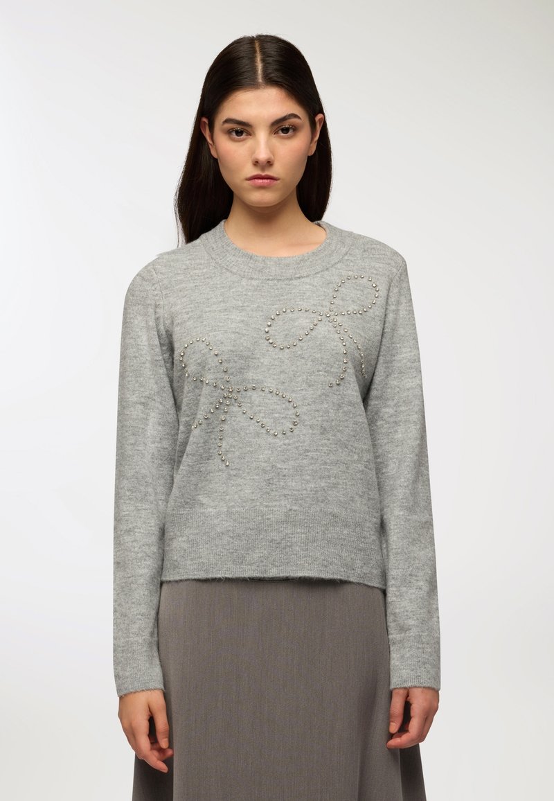 Donna che indossa un maglione grigio chiaro con decorazioni di perline a forma di fiocco, abbinato a una gonna grigia coordinata, su uno sfondo semplice.