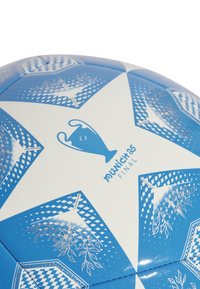 Balionas futbolui baltos ir mėlynos spalvų, su žvaigždės formos skydais, UEFA trofėjaus grafika ir lapų raštais blizgančioje tekstūroje.