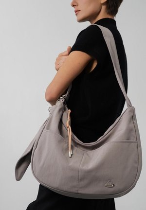 Femme en haut noir sans manches portant un grand sac à bandoulière gris clair avec une sangle réglable et un petit charm décoratif.