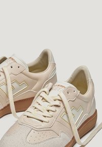 Baskets en cuir beige avec des accents texturés, présentant des coutures jaunes, une semelle en caoutchouc et des lacets tissés reposant lâchement.