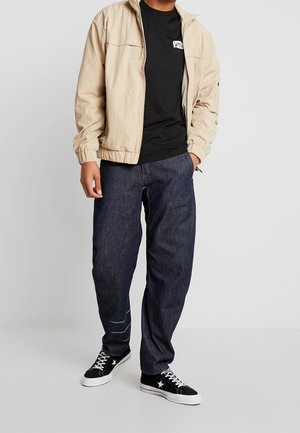 Mann trägt eine beigefarbene Jacke über einem schwarzen Vans-T-Shirt, dunkelblaue Jeans und schwarze Sneaker mit weißem Stern und weißen Schnürsenkeln, steht vor einem grauen Hintergrund.