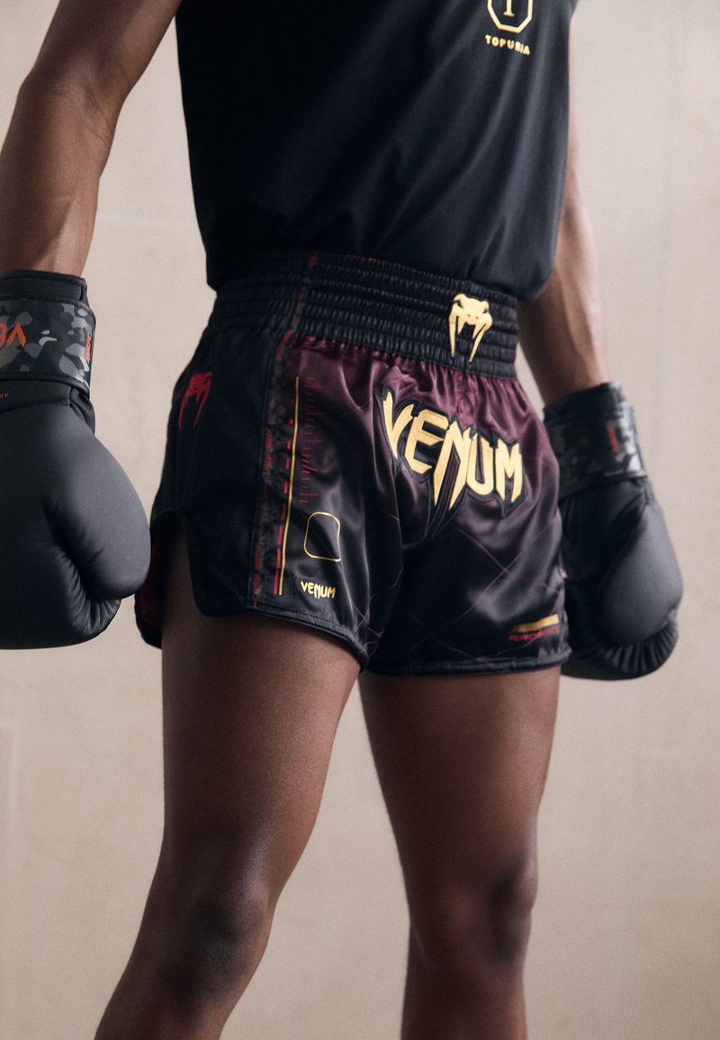 Črne in bordo boksarske hlače z zlatim logotipom "VENUM"; elastičen pas, stranske razpoke in kontrastni rdeči in rumeni detajli. Predstavljene so črne rokavice.