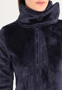 Donna che indossa una giacca a zip in pile blu navy con un colletto alto e ampio e un patch logo testurizzato sul petto.