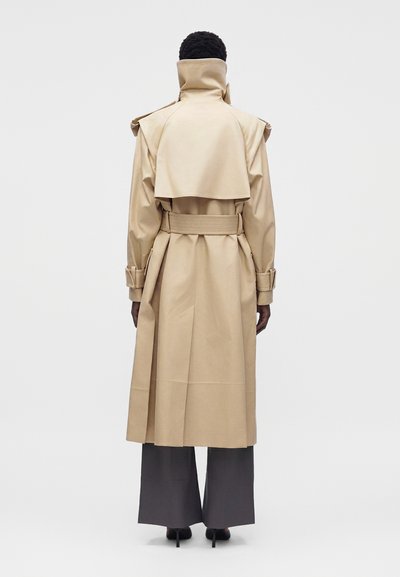 Personne portant un trench-coat beige avec un col montant et une ceinture, associé à un pantalon large gris foncé et des talons hauts noirs, debout de dos.