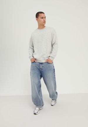 Sudadera gris de corte relajado, combinada con jeans de mezclilla azul de pierna ancha. Zapatillas deportivas plateadas completan el atuendo. Fondo liso.