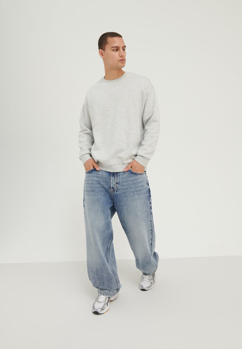 Grå sweatshirt i avslappet passform, kombinert med vide, blå denimjeans. Sølvfargede joggesko fullfører antrekket. Uspesifisert bakgrunn.