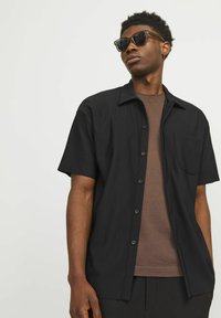 Black short-sleeve shirt with a textured finish, button front, and patch pocket, worn over a brown t-shirt. Model wears dark sunglasses.