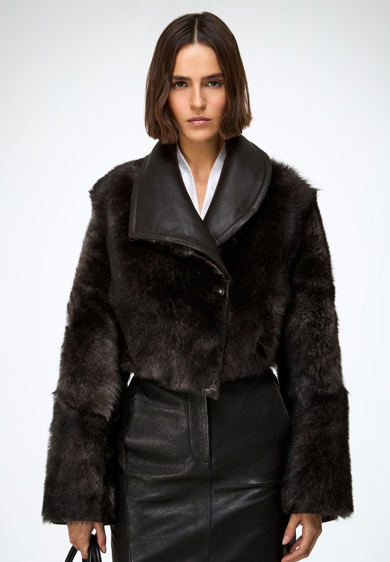 Kratka jakna od umjetnog krzna s crnim kožnim ovratnikom, teksturirane površine i širokih rukava, uparena s elegantnom crnom kožnom suknjom.