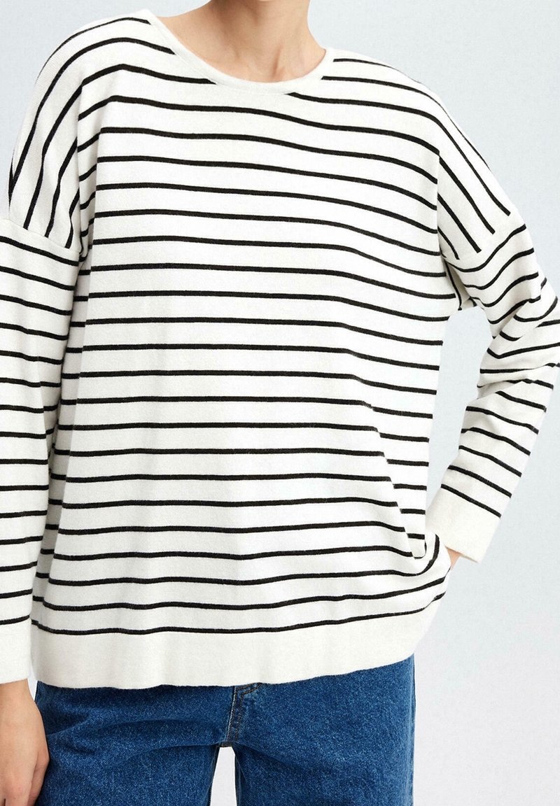 Person trägt ein weißes Langarmshirt mit dünnen schwarzen horizontalen Streifen und blaue Jeans, die Hand im Tasch, vor einem hellen Hintergrund.