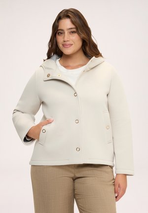 Frau mit welligem braunem Haar, die eine hellbeige Kapuzenjacke und khakifarbene Hose trägt, lächelt mit einer Hand in der Jackentasche.