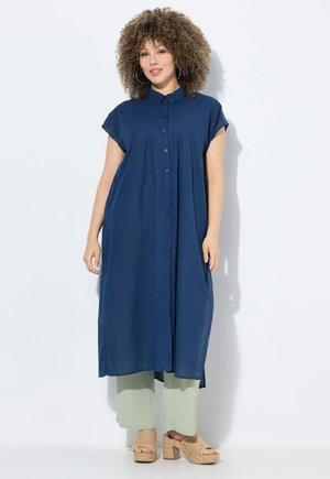 Abito blu navy con bottoni e maniche corte a cappuccio, con tasche laterali, indossato sopra pantaloni larghi verde chiaro; sandali testurizzati completano il look.