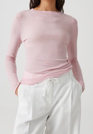 Femme portant un haut à manches longues rose clair rentré dans un pantalon blanc à cordon, les mains dans les poches, sur un fond clair uni.