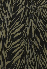 Olivově zelená látka s černým vzorem pruhů zebry, s dvěma černými knoflíky u výstřihu a texturovaným povrchem.