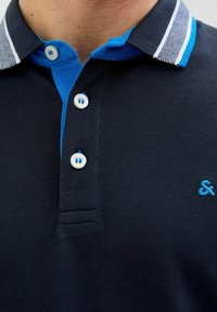 Marineblauwe polo met een contrasterende blauwe kraag, voorzien van drie knopen en een klein geborduurd logo in lichtblauw op de borst.