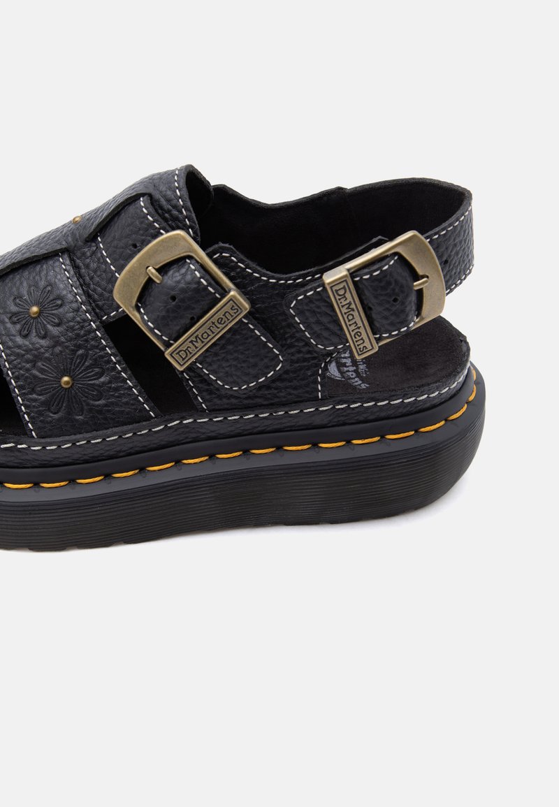 Schwarze Plateau-Sandale aus Leder mit floraler Prägung, zwei Messingschnallen mit der Aufschrift "Dr. Martens" und gelber Naht an der Sohle.