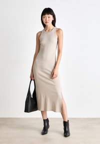Rochie midi râsucită bej, cu guler rotund și fante pe lateral, asortată cu o geantă tote neagră și cizme până la gleznă, pe un fundal neutru.