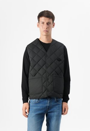 Ung man i en svart, vadderad väst över en långärmad skjorta, kombinerad med blå jeans, stående mot en enkel vit bakgrund.