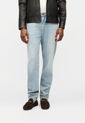 Άνδρας που φοράει φωτεινά μπλε τζιν, σκούρα καφέ μοκασίνια, λευκό μπλουζάκι και μαύρο δερμάτινο μπουφάν, στέκεται μπροστά σε απλό λευκό φόντο.