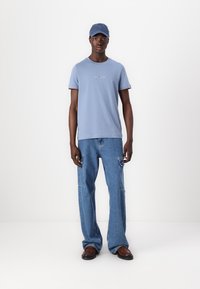 Light blue short-sleeve T-shirt with a contrasting neckline, paired with baggy light blue jeans featuring side pockets and brown shoes. Blue cap.