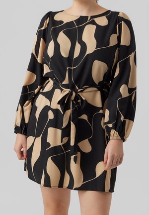 Kobieta w czarnej sukience z beżowymi abstrakcyjnymi wzorami, z długimi rękawami, elastycznymi mankietami i paskiem wiązanym w talii, stojąca na jednolitym tle.
