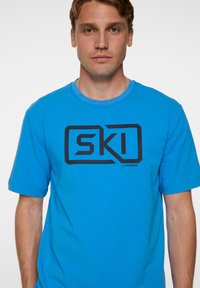 Kirkas sininen puuvillainen t-paita, jossa on rohkea musta "SKI" -logo, lyhyet hihat ja pyöreä kaula-aukko. sileä pinta ja rennon istuvuuden.