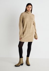 Beige kötött garbó ruha bordázott ujjal, fekete harisnyával és taupe bokacsizmával párosítva. Sima textúra és laza fazon.