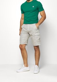 Grön bomullströja med ett litet emblem och beige cargobyxor med sidofickor, bärs med vita sneakers mot en plain bakgrund.