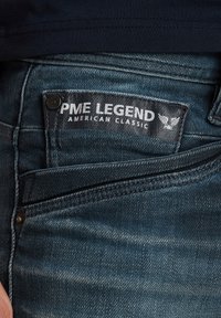 Plave traperice s tamnim kožnim zakrpama na kojima je uvezano "PME Legend American Classic", ukrašene metalnim gumbom blizu džepa.