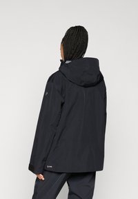 Μαύρο αδιάβροχο μπουφάν με κουκούλα, διαθέτει μοντέρνο, χαλαρό σχέδιο, τσιμέντο velcro στα μανίκια και λογότυπο κοντά στο στρίφωμα.