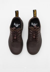 Brown leather lace-up shoes with a rounded toe, textured surface, reinforced stitching, and a yellow pull tab at the heel. Dr. Martens branding inside.