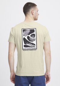 Lys beige t-skjorte med en svart grafisk design på baksiden med blader og teksten "TILLAT DEG SELV Å VOKSE" og "UTSTRÅLE POSITIVITET."